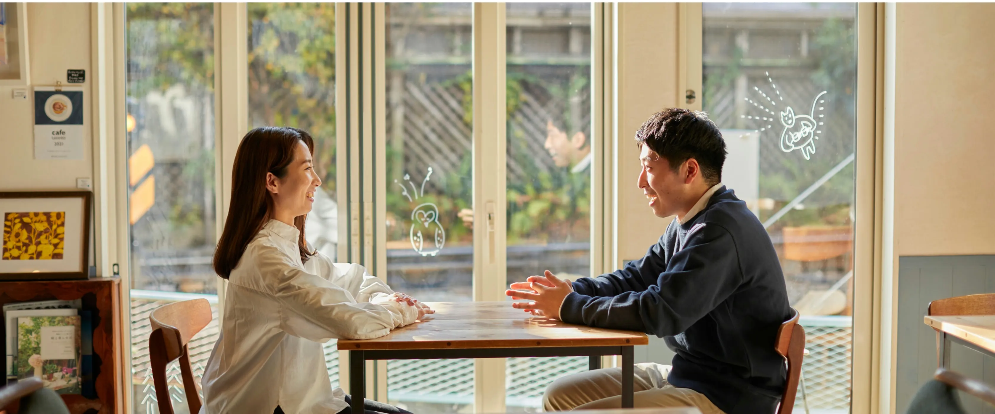 明るいカフェの窓際で、テーブルをはさんで笑顔で話す男女